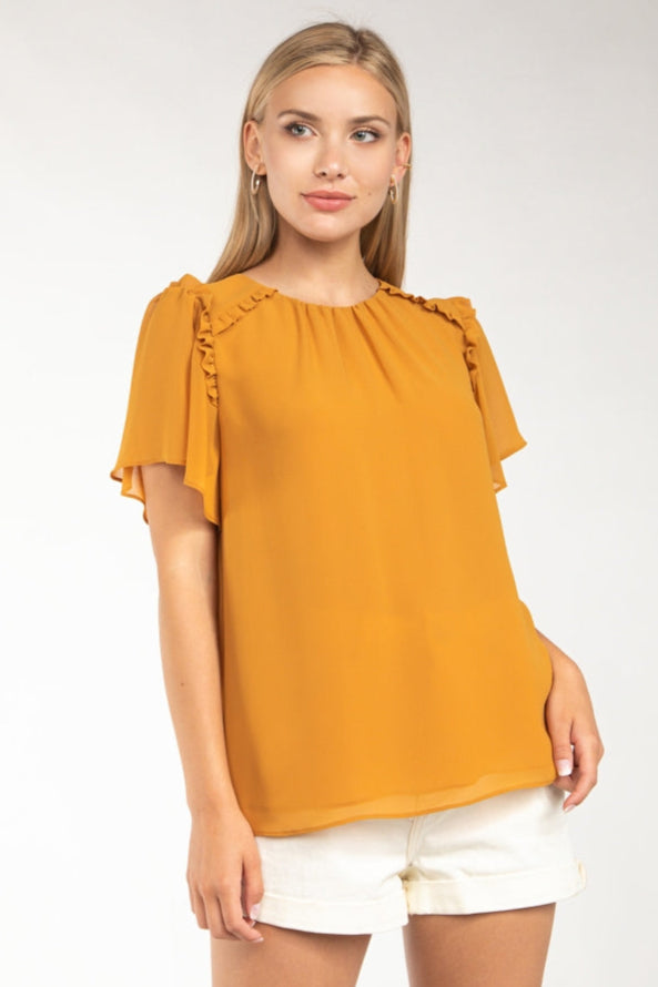 A female model wearing a short sleeve blouse with ruffle trim and paired with white shorts.