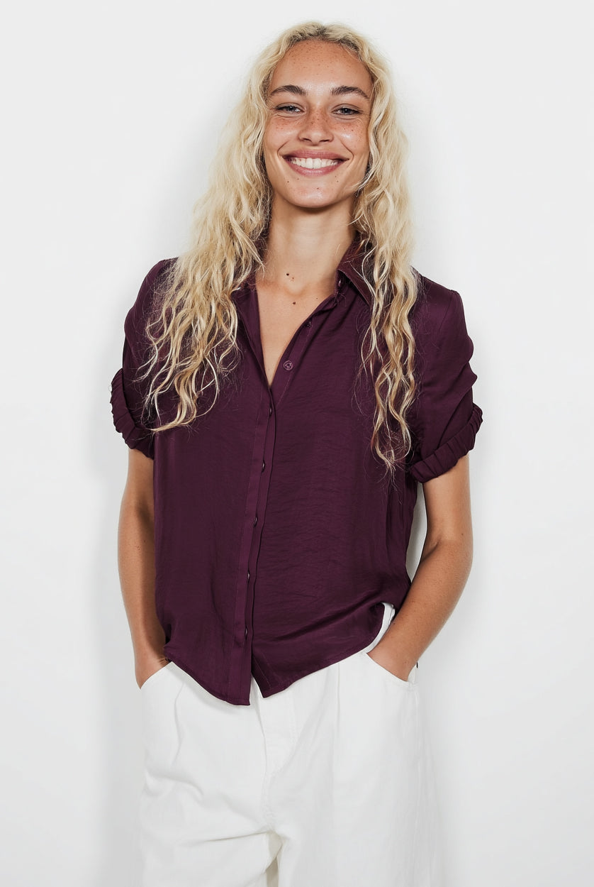 Woman wearing a dark purple shirt and white pants against a plain background