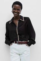 Woman wearing a black blouse with white trim and white pants against a plain background